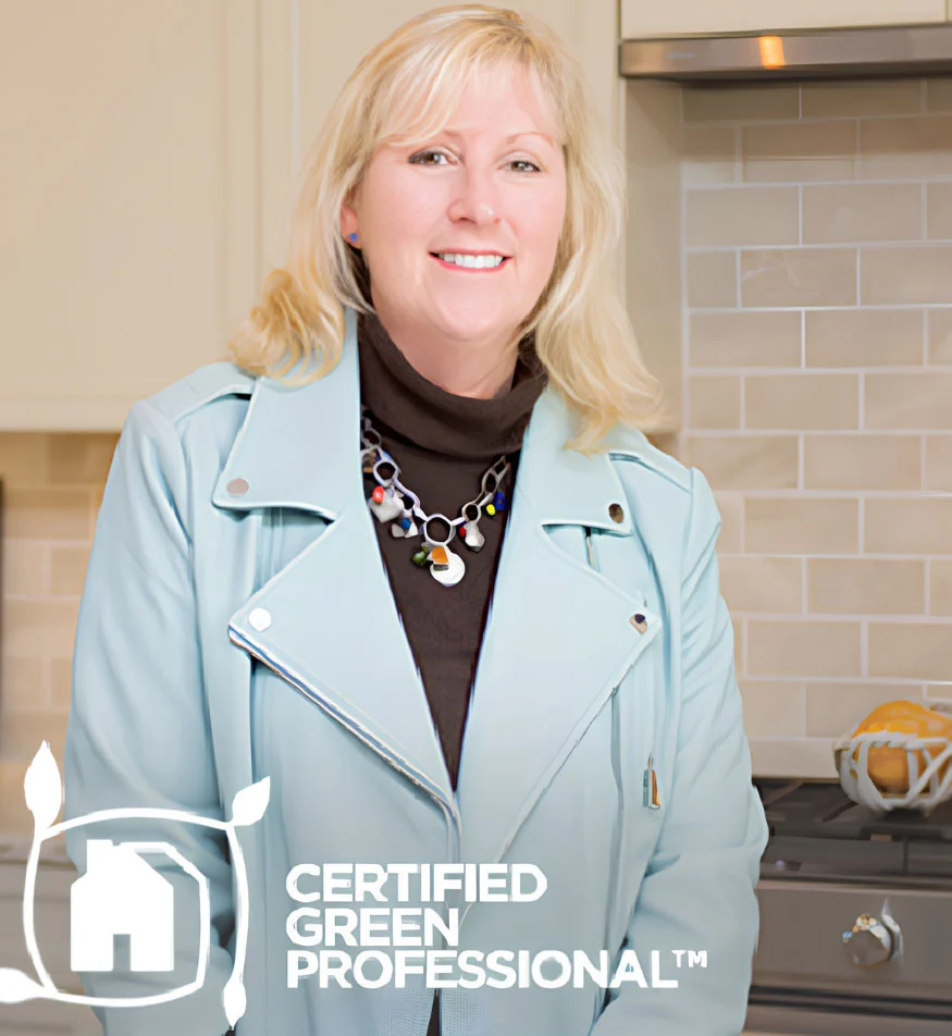 Smiling woman in a light blue jacket with "Certified Green Professional" logo, standing in a modern kitchen, representing eco-friendly home builder.