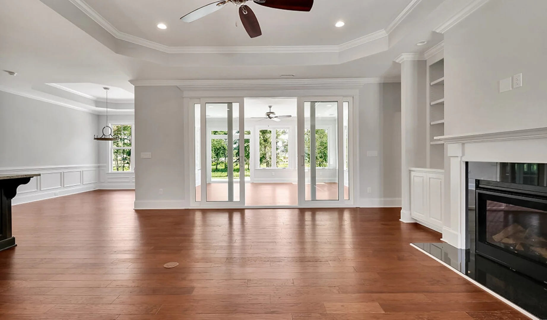 Spacious living area with hardwood floors, ceiling fan, modern fireplace, and sliding glass doors leading to a bright sunroom. Ideal for home builders