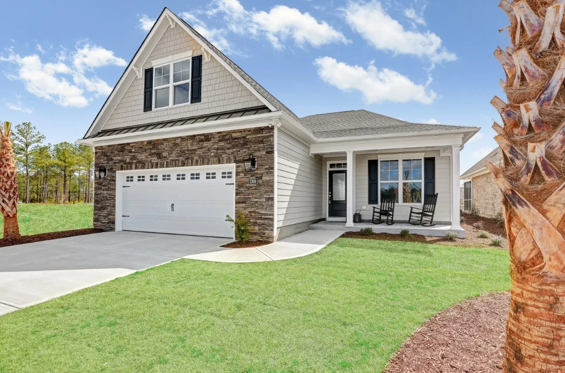 Modern suburban house with stone facade, white garage, and cozy porch. Ideal design by home builder amidst lush greenery under a clear sky.