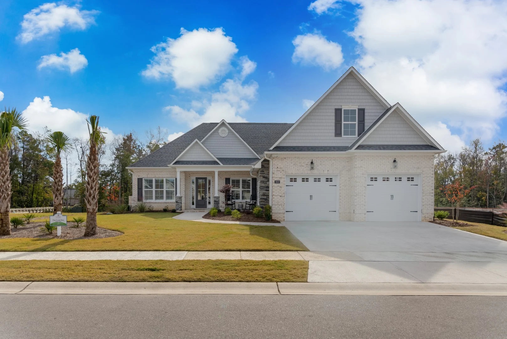 Modern single-story suburban home with gray shingles, two-car garage, manicured lawn, and palm trees, showcasing a home builder's design.