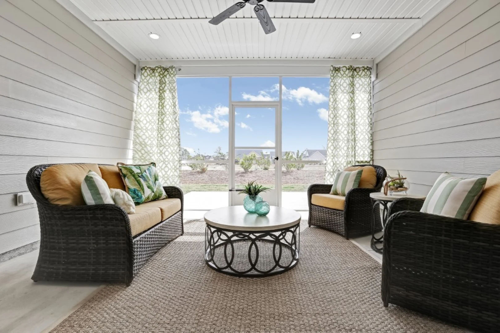 Cozy sunroom with rattan chairs, cushions, round table, green curtains, ceiling fan, built by a home builder. Bright view through large window.