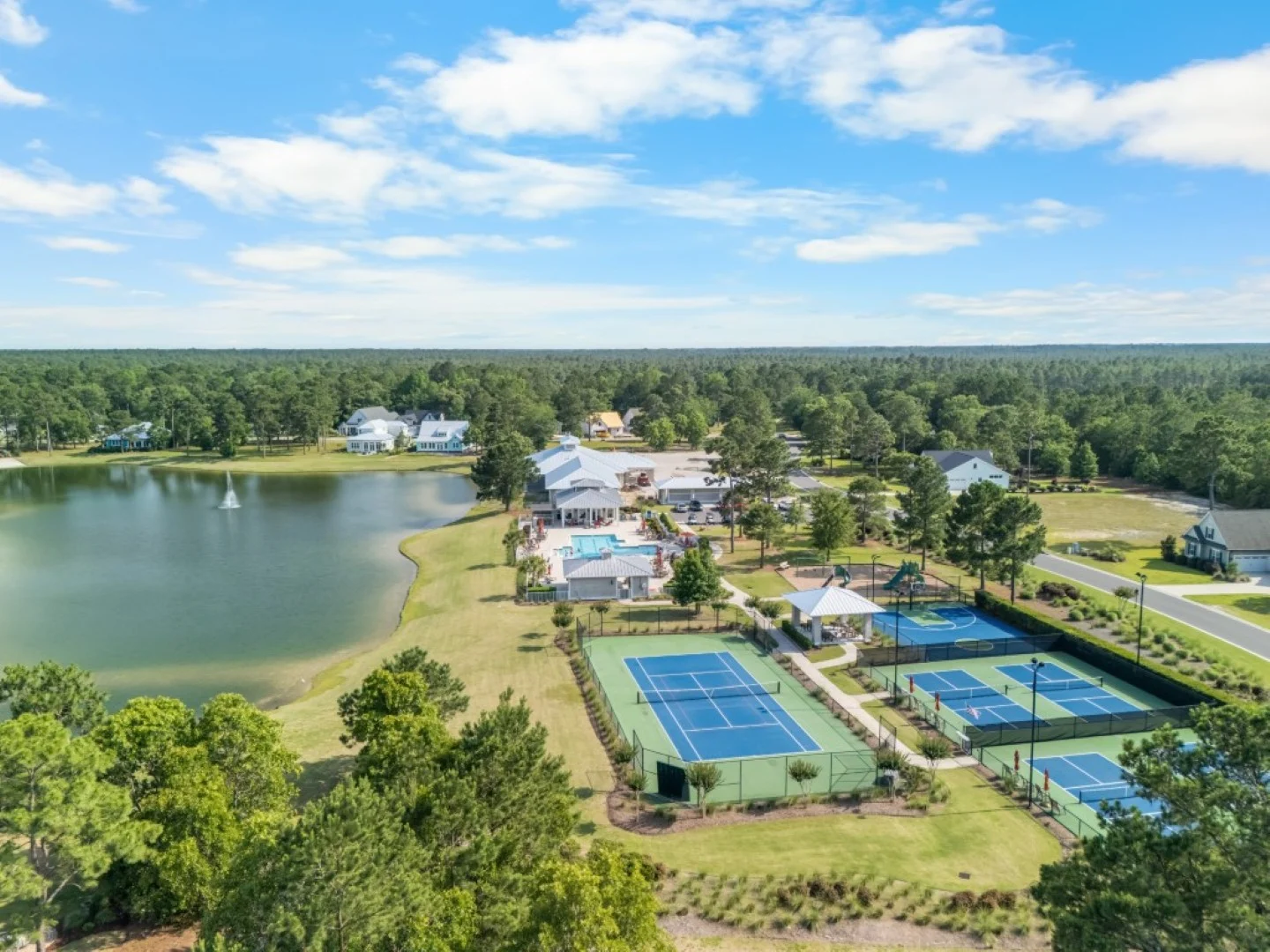 Aerial view of a community with a lake, tennis courts, pool, and homes surrounded by trees, showcasing a home builder's development.