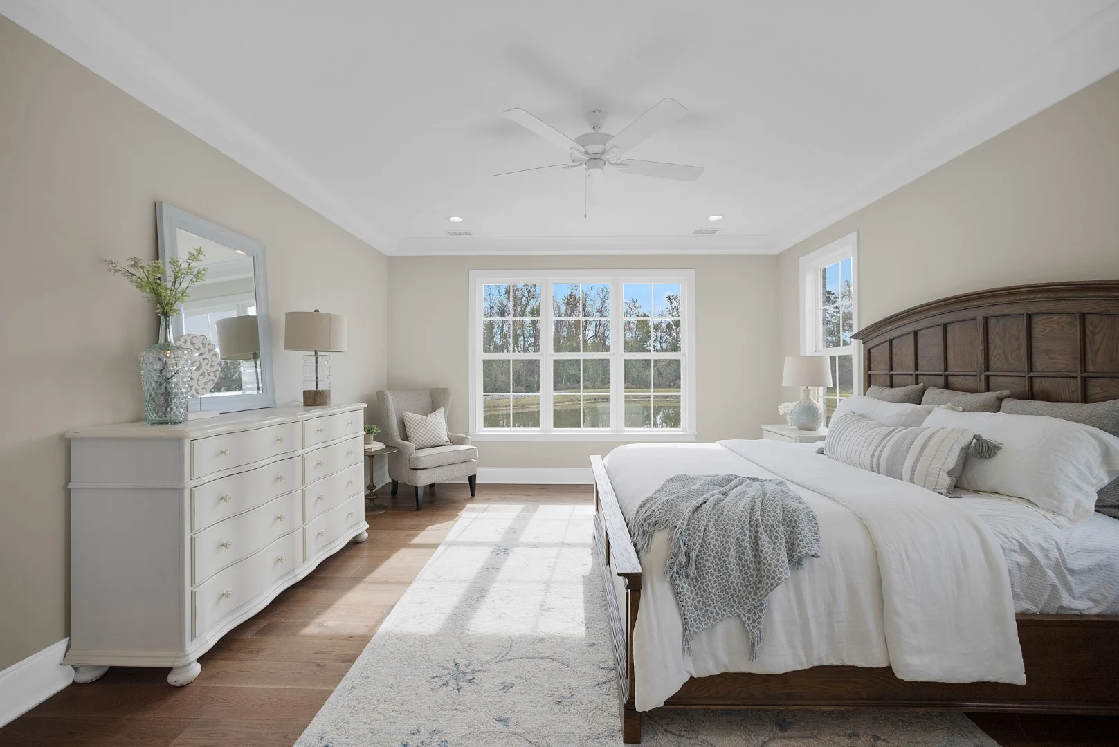 Bright bedroom by a home builder, featuring a large bed, wooden floor, and cozy armchair by sunlit windows with a garden view.
